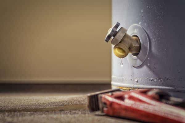 A close-up of a water heater valve with a small leak, showing water dripping onto the floor. A red pipe wrench lies on the ground nearby, hinting at an upcoming water heater replacement that may require a permit for water heater work. | High 5 Plumbing, Heating & Cooling