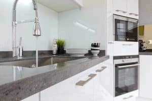 Modern kitchen with a gray countertop, stainless steel sink, and chrome pull-down faucet. Stacked white plates and bowls sit by small plants—perfect for those looking to improve water quality with stylish fixtures in white cabinetry. | High 5 Plumbing, Heating & Cooling