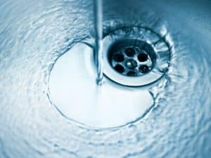 A close-up image of water flowing from a tap into a metal sink, tinted blue, with bubbles and ripples swirling around the drain—perfectly illustrating the clarity achieved with a water filtration purifier. | High 5 Plumbing, Heating & Cooling