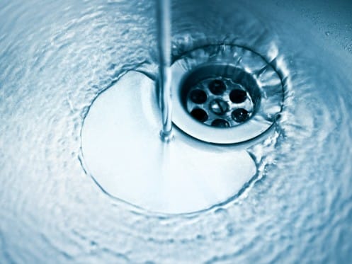 A close-up image of water flowing from a tap into a metal sink, tinted blue, with bubbles and ripples swirling around the drain—perfectly illustrating the clarity achieved with a water filtration purifier. | High 5 Plumbing, Heating & Cooling
