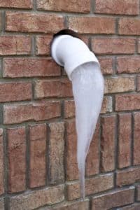 A PVC pipe protrudes from a brick wall in Denver, CO, with a large, pointed icicle hanging from its opening—a reminder to follow winter plumbing tips to protect pipes from freezing temperatures. | High 5 Plumbing, Heating & Cooling