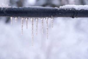 A row of icicles hangs from a snow-covered black metal pipe, with a blurred snowy background—a reminder of the importance of safely thawing pipes to prevent plumbing disasters during harsh winter weather. | High 5 Plumbing, Heating & Cooling