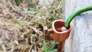 A close-up view of a copper grounding rod with a green and yellow ground wire attached by a metal clamp, positioned next to a concrete surface and surrounded by grass, highlights the importance of electrical grounding for safety. | High 5 Plumbing, Heating & Cooling