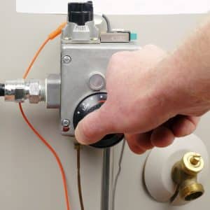 A hand turns the temperature control dial on a water heater valve, highlighting water heater energy usage, with "Pilot" labeled above and pipes attached to the unit. | High 5 Plumbing, Heating & Cooling