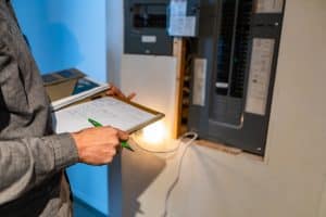 A person holding a clipboard and pen stands near an open residential electrical panel, inspecting or recording information from the circuit breakers, possibly assessing when to upgrade electrical panel components. | High 5 Plumbing, Heating & Cooling