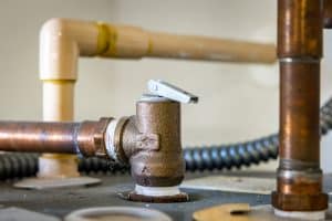 Close-up of a water heater pressure relief valve attached to a copper pipe, with additional plastic and metal pipes in the background. The valve is installed on a flat metal surface. | High 5 Plumbing, Heating & Cooling