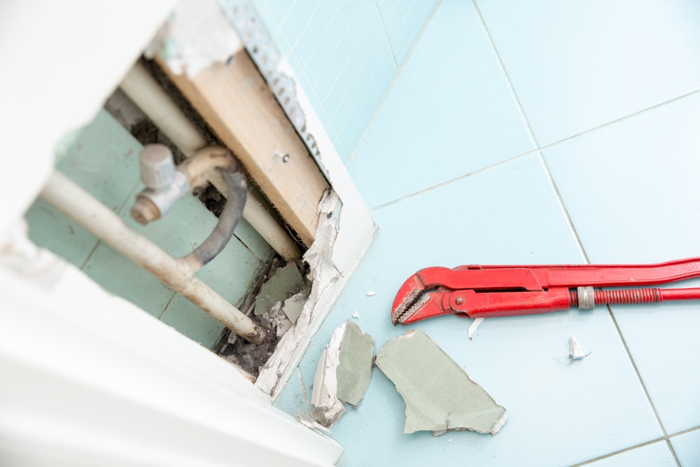 A red pipe wrench lies on a tiled floor next to broken drywall, exposing pipes inside a wall—evidence of a hidden plumbing leak recently uncovered during plumbing leak detection. | High 5 Plumbing, Heating & Cooling